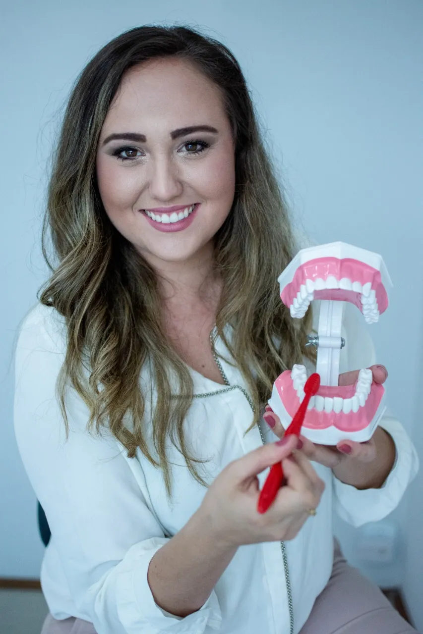Dentista da clínica sorrindo, segurando um modelo de arcada dentária e uma escova